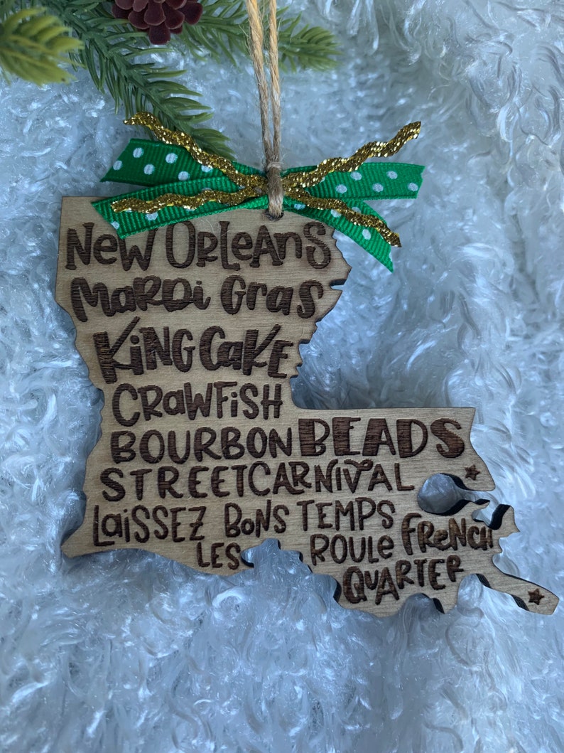 May include: Wooden ornament shaped like the state of Louisiana with the words "New Orleans Mardi Gras King Cake Crawfish Bourbon Beads Streetcarnival Laissez Les Bons Temps Roule French Quarter" written on it. The ornament is decorated with a green and gold ribbon.