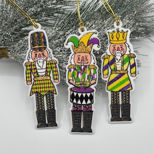 May include: Three Mardi Gras-themed nutcracker ornaments. Each ornament features a different design, including a hat or crown, and is decorated in green, purple, and gold. They have gold hanging strings and black boots.