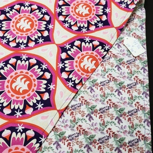 Può includere: Un tessuto bianco con un motivo floreale rosa, arancione e viola da un lato e un tessuto bianco con un motivo floreale verde e viola dall'altro lato. Il tessuto è piegato a metà e un'etichetta è attaccata all'angolo che recita "11 1/2 x 15 1/2"