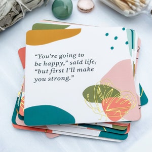 May include: A stack of square cards with a white background and colorful abstract shapes in coral, teal, and mustard yellow. The cards feature the quote: "You're going to be happy," said life, "but first I'll make you strong." A gold butterfly design is also present.