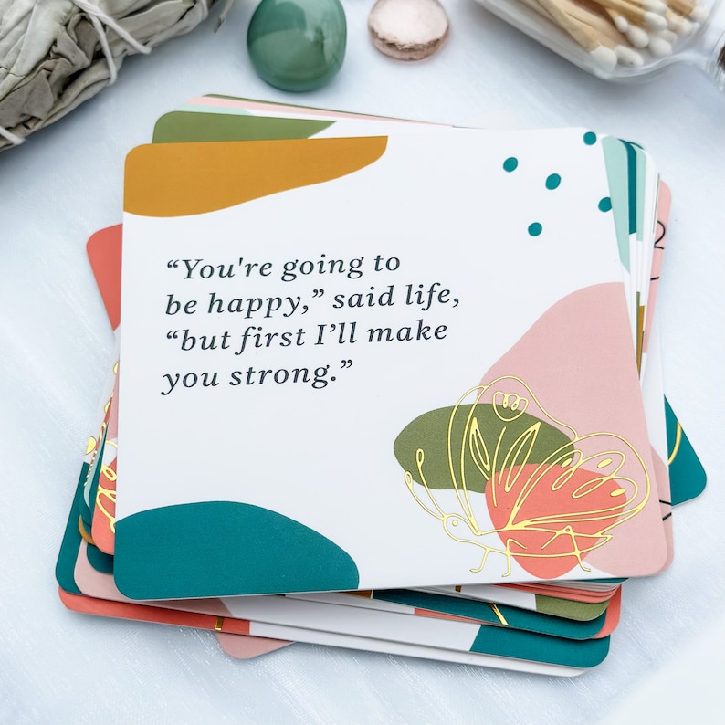May include: A stack of square cards with a white background and colorful abstract shapes in coral, teal, and mustard yellow. The cards feature the quote: "You're going to be happy," said life, "but first I'll make you strong." A gold butterfly design is also present.