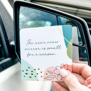 May include: A square card held near a car mirror features the text "The rear view mirror is small for a reason." The card has a white background with abstract shapes in teal, peach, and green, plus gold dots and a floral design.