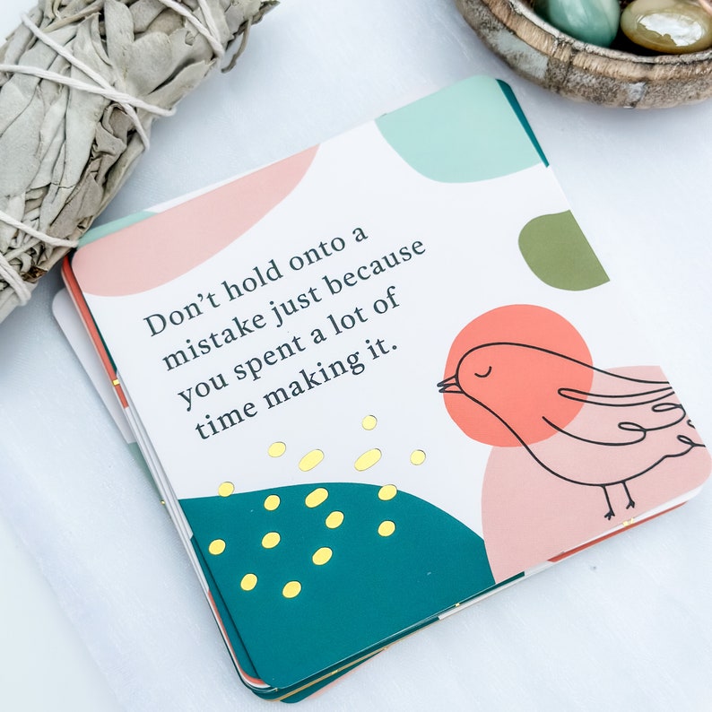 May include: A stack of square cards with a colorful design and the text "Don't hold onto a mistake just because you spent a lot of time making it." The cards feature a bird illustration and gold accents.