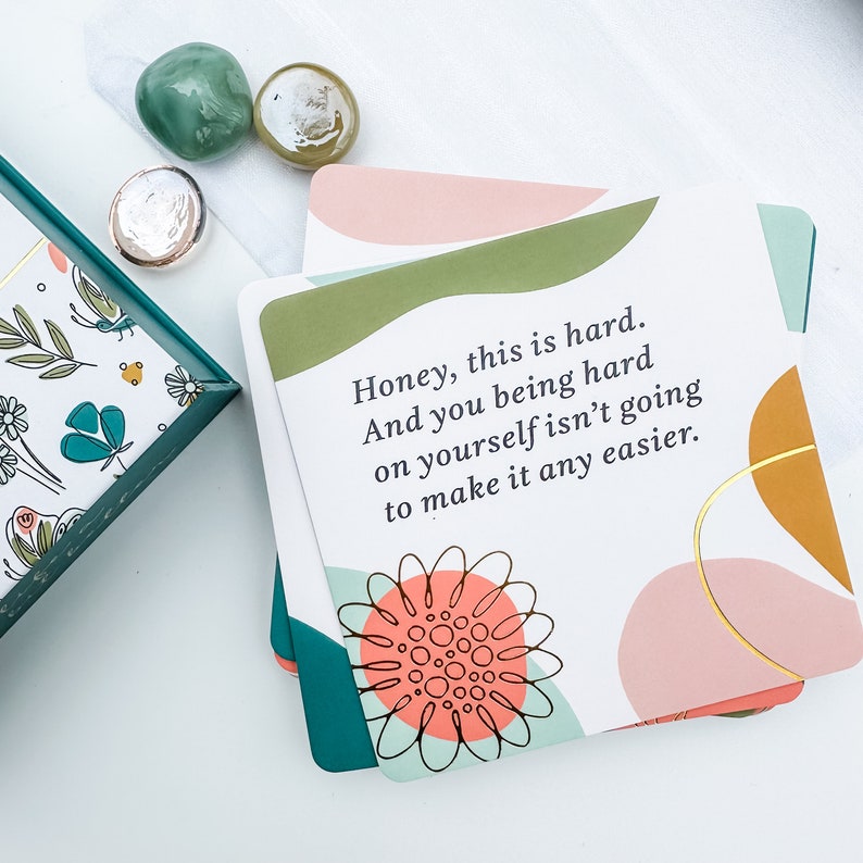 May include: A stack of white cards with a floral design and the text "Honey, this is hard. And you being hard on yourself isn't going to make it any easier." The cards have a teal border and are decorated with abstract shapes and a flower.