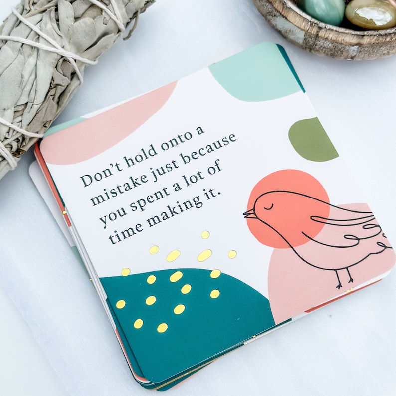 May include: A stack of square cards with a colorful design and the text "Don't hold onto a mistake just because you spent a lot of time making it." The cards feature a bird illustration and gold accents.
