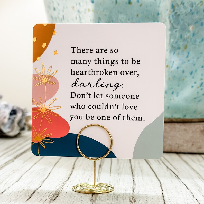 May include: A square card with a gold wire holder features the text "There are so many things to be heartbroken over, darling. Don't let someone who couldn't love you be one of them." The card has abstract art.