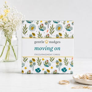 以下が含まれることがあります： gentle nudges moving onというタイトルの励ましカードが入った正方形の箱。箱は白い背景で、ティールブルーの蝶、黄色い花、緑の葉が繰り返される模様が描かれています。テキストは中央にティールブルーとダークグレーで配置されています。