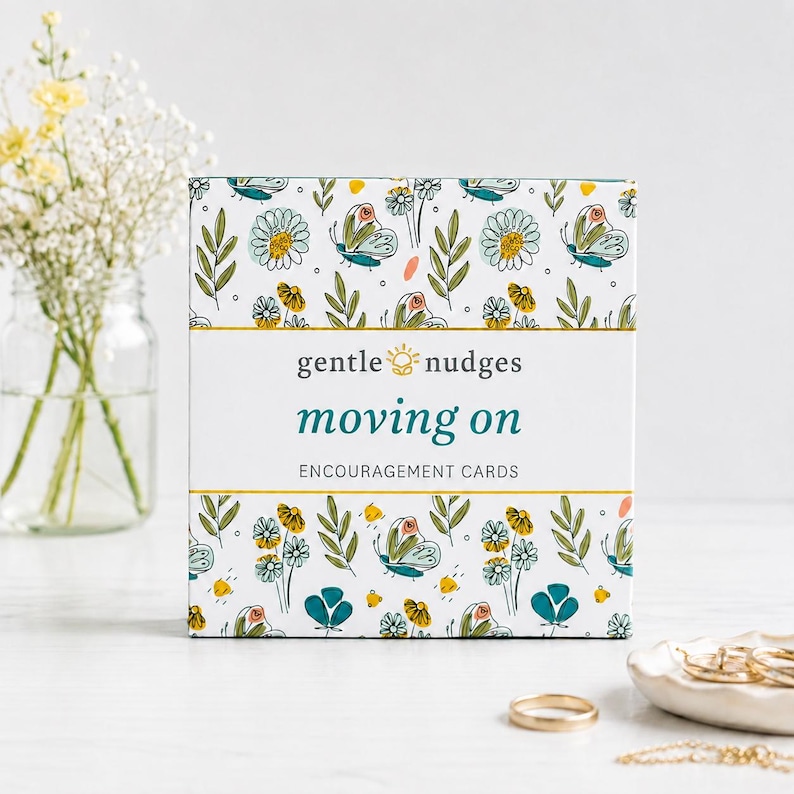 以下が含まれることがあります： gentle nudges moving onというタイトルの励ましカードが入った正方形の箱。箱は白い背景で、ティールブルーの蝶、黄色い花、緑の葉が繰り返される模様が描かれています。テキストは中央にティールブルーとダークグレーで配置されています。