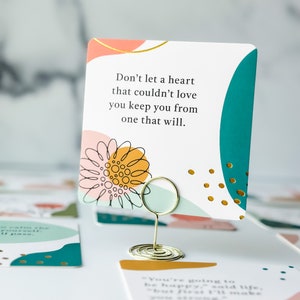 May include: A square card with a gold wire holder, featuring the text "Don't let a heart that couldn't love you keep you from one that will." The card has a teal, orange, and pink design with gold accents. Other cards are visible in the background.