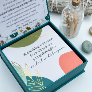 May include: A teal box with a set of cards featuring the text "Something will grow from all you are going through... and it will be you." The box is open, revealing the cards. A glass bottle with matches and decorative stones are in the background.