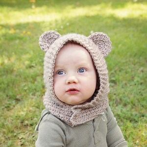 Peut inclure: Un bébé portant un bonnet en crochet marron en forme d'ours avec des oreilles, un t-shirt à manches longues en tricot gaufré vert et un pantalon camouflage.