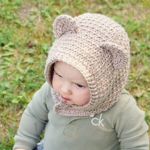 Peut inclure: Un bébé portant un bonnet en crochet beige avec des oreilles d'ours. Le bonnet est de conception en crochet et couvre les oreilles du bébé.