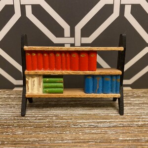 May include: A miniature wooden bookshelf with two shelves, painted black, holding a collection of miniature books with red, green, and blue covers.