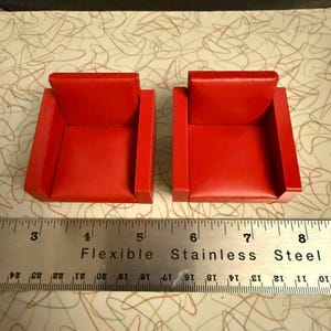 May include: Two miniature red armchairs. Each chair has a square seat and back, with armrests. The chairs are placed next to a ruler that measures in inches. The chairs are likely for a dollhouse or model display.