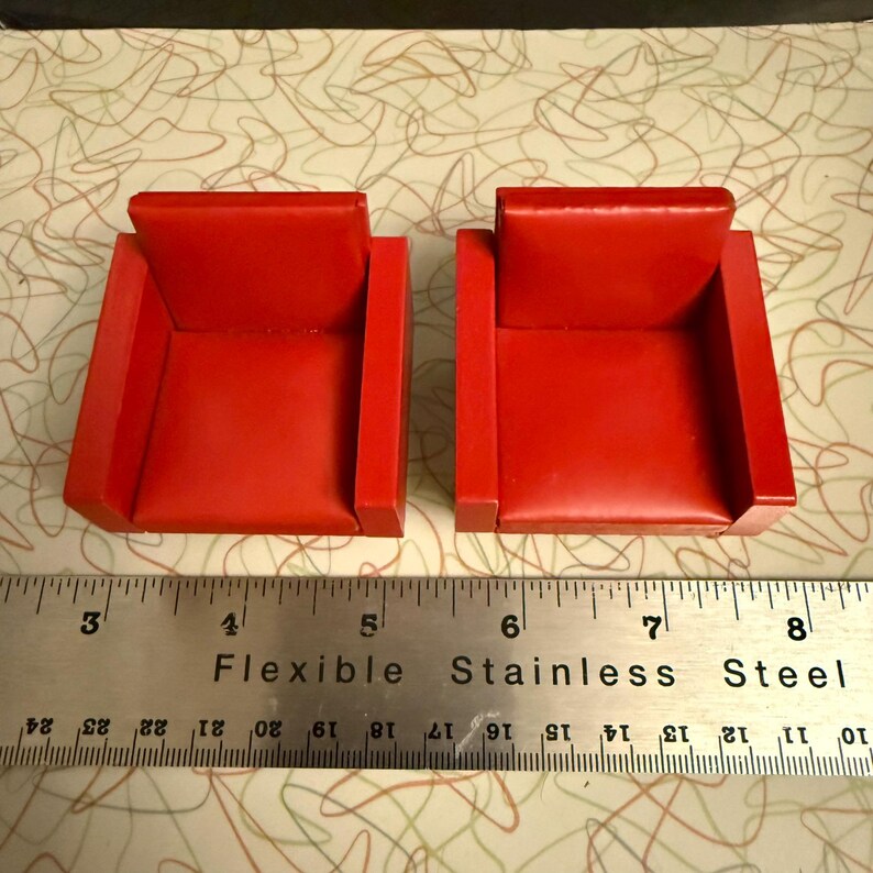 May include: Two miniature red armchairs. Each chair has a square seat and back, with armrests. The chairs are placed next to a ruler that measures in inches. The chairs are likely for a dollhouse or model display.