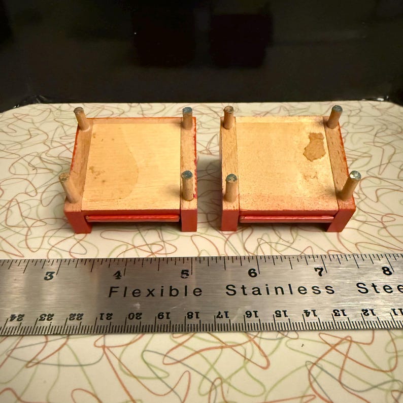 May include: Two miniature wooden beds with red painted frames and natural wood platforms. Each bed has four cylindrical legs with silver-colored tips. A metal ruler with the text "Flexible Stainless Steel" is placed near the beds.