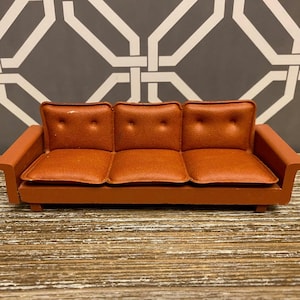 May include: A miniature orange sofa with three seat cushions and button detailing. The sofa has a low profile with armrests on each side. The sofa is made of a smooth material and sits on a wooden base.