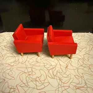 May include: Two miniature red armchairs with light wood legs. The chairs have a mid-century modern design, with a boxy shape and a simple, clean aesthetic. The chairs are placed on a patterned surface with a black background.