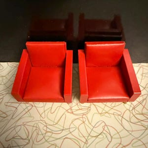 May include: Two small, red armchairs with a boxy design. The chairs have a smooth, glossy finish and are set against a patterned background. The armchairs are a vibrant red color.