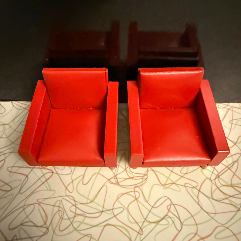 May include: Two small, red armchairs with a boxy design. The chairs have a smooth, glossy finish and are set against a patterned background. The armchairs are a vibrant red color.