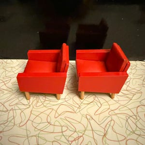 May include: Two miniature red armchairs with light wood legs are displayed. The chairs have a simple, boxy design with a low back and armrests. The chairs are set against a patterned background.