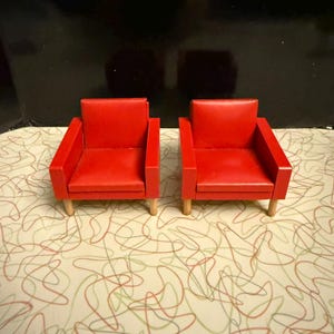 May include: Two miniature red armchairs with wooden legs. The chairs have a simple, boxy design with a smooth, glossy finish. The background is a patterned surface with a black backdrop.