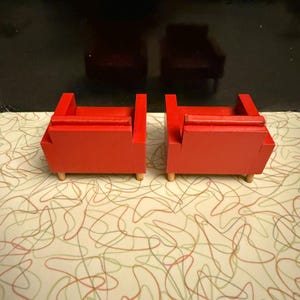 May include: Two small, bright red wooden chairs with a mid-century modern design. Each chair has a rectangular seat and back, with a small armrest on each side. The chairs are supported by four short, light-colored wooden legs.