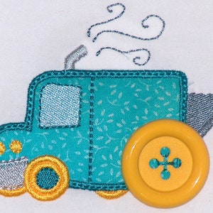 Könnte beinhalten: Ein türkisfarbenes Spielzeugauto mit einem gelben Knopf an der Seite. Das Auto hat einen silbernen Auspuff und ein weißes Fenster. Das Auto ist mit einem weißen Blumenmuster bestickt.