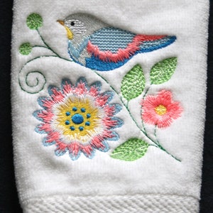Pode incluir: Toalha de mão branca bordada com um pássaro azul empoleirado em um caule verde com flores rosa e amarelas.