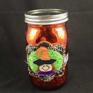 May include: A metallic orange glass jar with a silver screw-top lid. An appliqued fabric patch is attached to the front, featuring a cartoon witch with a pointed orange hat, a black brim, and a purple collar on a green and black spiderweb background.