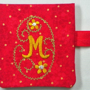 Op de afbeelding: Rood stoffen item met gouden stippen en een geel geborduurde letter "M". De letter is omgeven door groene ranken en gele bloemen. Een rode elastische band is aan de zijkant bevestigd.