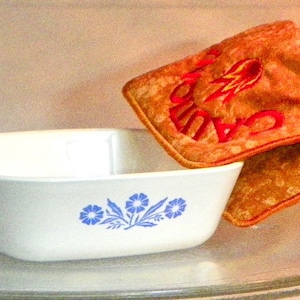 May include: A white ceramic baking dish with a blue floral design is being removed from a microwave using a brown oven mitt with the text "Care" embroidered in red.