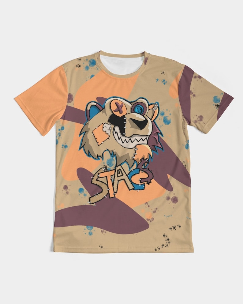 Op de afbeelding: Een beige T-shirt met een cartoon leeuwenontwerp. De leeuw heeft gestikte patches, een ooglapje en een knoog. Het woord "STA" staat eronder. Het T-shirt heeft een oranje en paars abstract ontwerp.