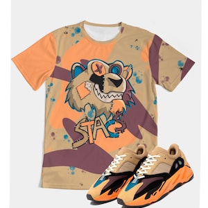 Op de afbeelding: Een beige t-shirt met een cartoon leeuw afbeelding en het woord "STAG" in oranje. Het shirt wordt gecombineerd met beige en oranje sneakers met zwarte en blauwe accenten. Het shirt heeft een camouflagepatroon.