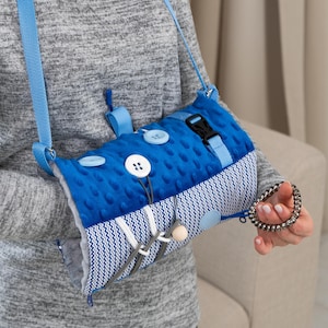 May include: A blue sensory bag with a textured surface, buttons, and a buckle. The bag has a blue strap and a gray and white patterned section. It is designed to provide tactile stimulation and sensory exploration.