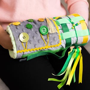 Puede incluir: Un juguete sensorial gris con diversas texturas y elementos. Tiene botones, cintas y una superficie suave y afelpada. El juguete está acentuado con detalles verdes, amarillos y blancos, diseñado para la exploración táctil y el juego.