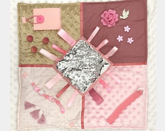 May include: A sensory activity square in shades of pink, tan, and mauve. Features include a sequined square, ribbons, buttons, a flower, and a beaded tassel. The square is designed with various textures and elements for tactile exploration.