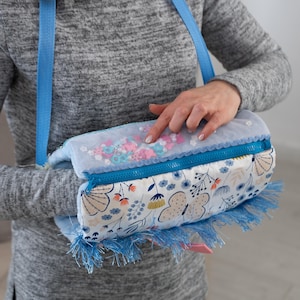 Puede incluir: Bolso bandolera con estampado floral azul y blanco, cremallera y correa azules. El bolso tiene una parte superior de malla llena de confeti rosa y azul. La parte inferior del bolso tiene flecos azules.