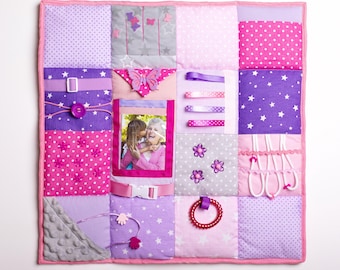 May include: A sensory activity board with a variety of textures and interactive elements. The board features a patchwork design in shades of pink, purple, and gray, with a photo frame, ribbons, and a beaded ring. The board is designed for tactile exploration.