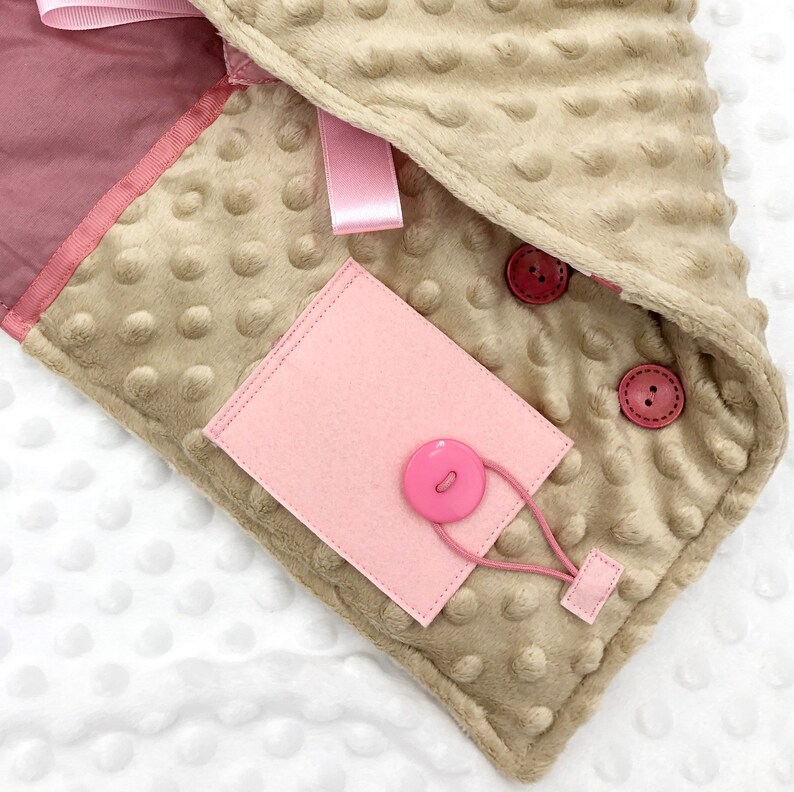 May include: A beige and pink baby blanket with a textured, dotted surface. The blanket features a pink felt tag with a button and elastic loop, along with two pink buttons and a pink ribbon. The blanket is made of soft material.