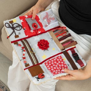 Pode incluir: Um quadro de atividades sensoriais com várias texturas e elementos interativos. Inclui um zíper, fitas, uma fivela, um botão e uma rosa de feltro. O quadro tem um fundo cinza com padrões de estrelas e detalhes em vermelho.