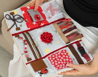 May include: A sensory activity board with various textures and interactive elements. Features include a zipper, ribbons, a buckle, a button, and a felt rose. The board has a gray background with star patterns and red accents.