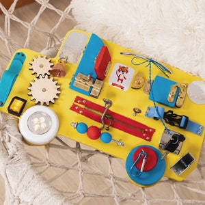 Busy board in the shape of track/bus with wheels and doors. The base is made of plywood. The thickness of the plywood is 10 mm (0.4 inches). Bright colors: yellow base, red and blue elements.