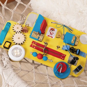 Busy board in the shape of track/bus with wheels and doors. The base is made of plywood. The thickness of the plywood is 10 mm (0.4 inches). Bright colors: yellow base, red and blue elements.