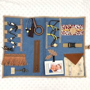 Puede incluir: Un tablero sensorial de mezclilla azul con varios elementos táctiles para el desarrollo de la motricidad fina. Incluye cordones, cremalleras, hebillas, un marco de fotos y telas texturizadas en marrón, beige y amarillo. Diseñado para la exploración sensorial.