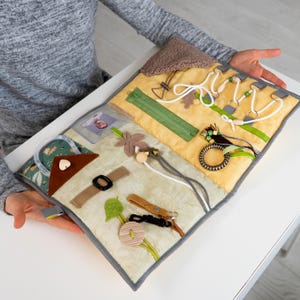 Puede incluir: Un tablero de actividades sensoriales con varias texturas y cierres. El tablero presenta una base de tela amarilla y verde claro con un borde gris. Incluye una cremallera, una hebilla y otros elementos interactivos.