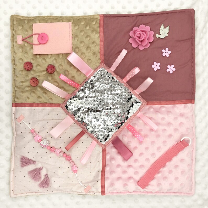 May include: A sensory activity square in shades of pink, tan, and mauve. Features include a sequined square, ribbons, buttons, a flower, and a beaded bracelet with tassels. The square is designed with various textures and elements.