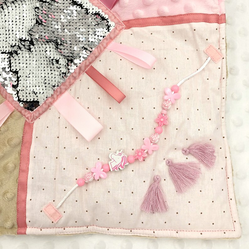 May include: A sensory activity square with various textures and embellishments. Features a section of reversible sequins, pink ribbons, and a beaded strand with a unicorn charm and flower accents. Includes pink tassels and a soft, dotted fabric.