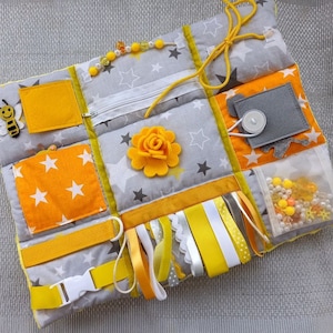 May include: A yellow and gray baby sensory activity mat with a zipper, a bee applique, a flower, ribbons, and a clear pocket filled with beads and small toys.