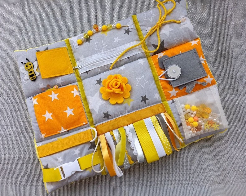 May include: A yellow and gray baby sensory activity mat with a zipper, a bee applique, a flower, ribbons, and a clear pocket filled with beads and small toys.